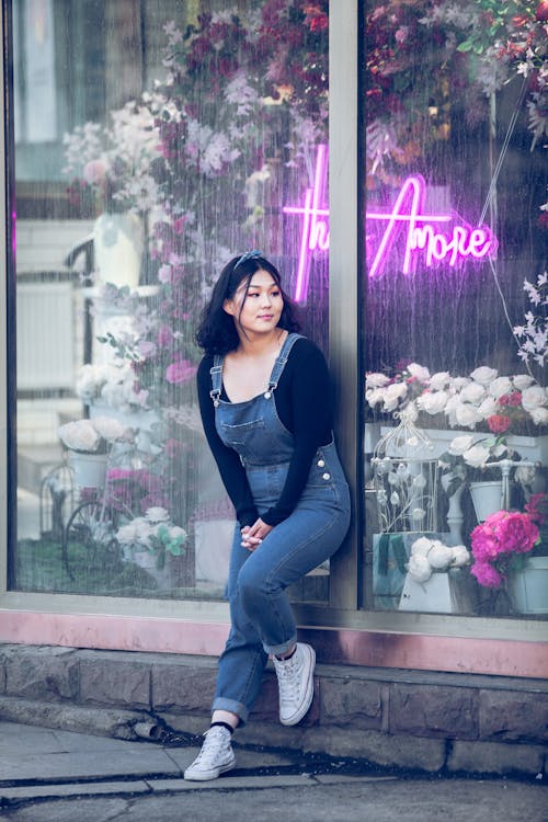 Content Asian female in denim jumpsuit standing near flower shop on city street and looking away