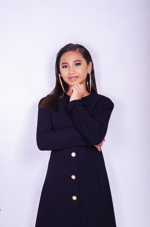 Free Young ethnic woman with dark long hair in trendy black coat and big earrings standing with crossed hands and touching chin while looking at camera on light lilac background Stock Photo