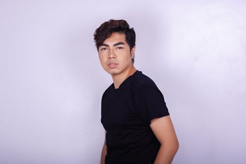 Serious young ethnic man in trendy clothes looking pensively at camera while standing against light lilac wall in modern studio