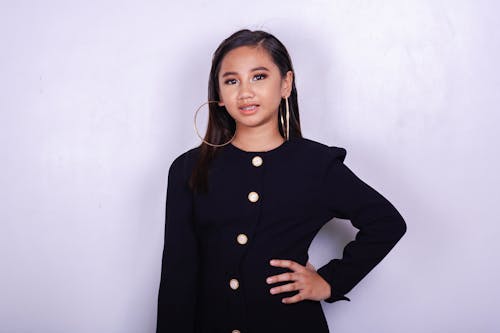 Positive young ethnic woman in trendy black clothes and big earrings standing with hand on waist and looking at camera against lilac wall