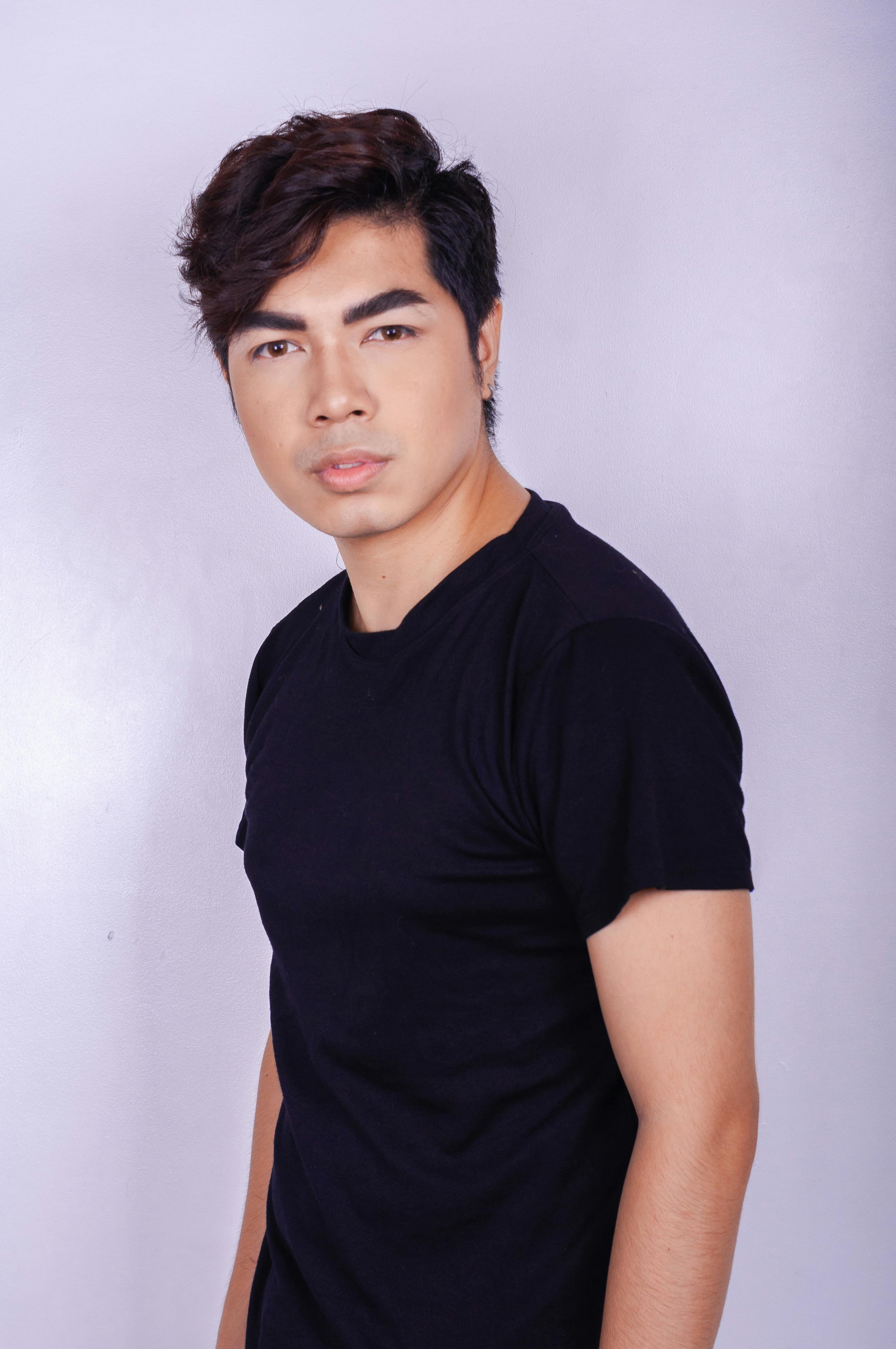 Confident young ethnic guy in trendy casual clothes looking seriously at camera while standing in modern studio against lilac wall