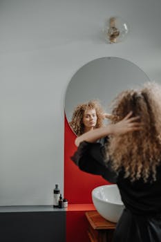 A young woman with curly hair styles herself in front of a modern bathroom mirror.