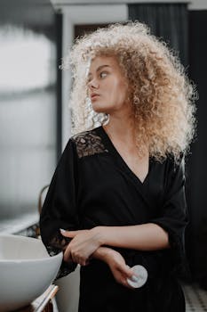 Portrait of a young woman with curly hair in a black robe, standing in a bathroom.