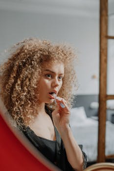 A young woman with curly hair brushes her teeth in front of a mirror.