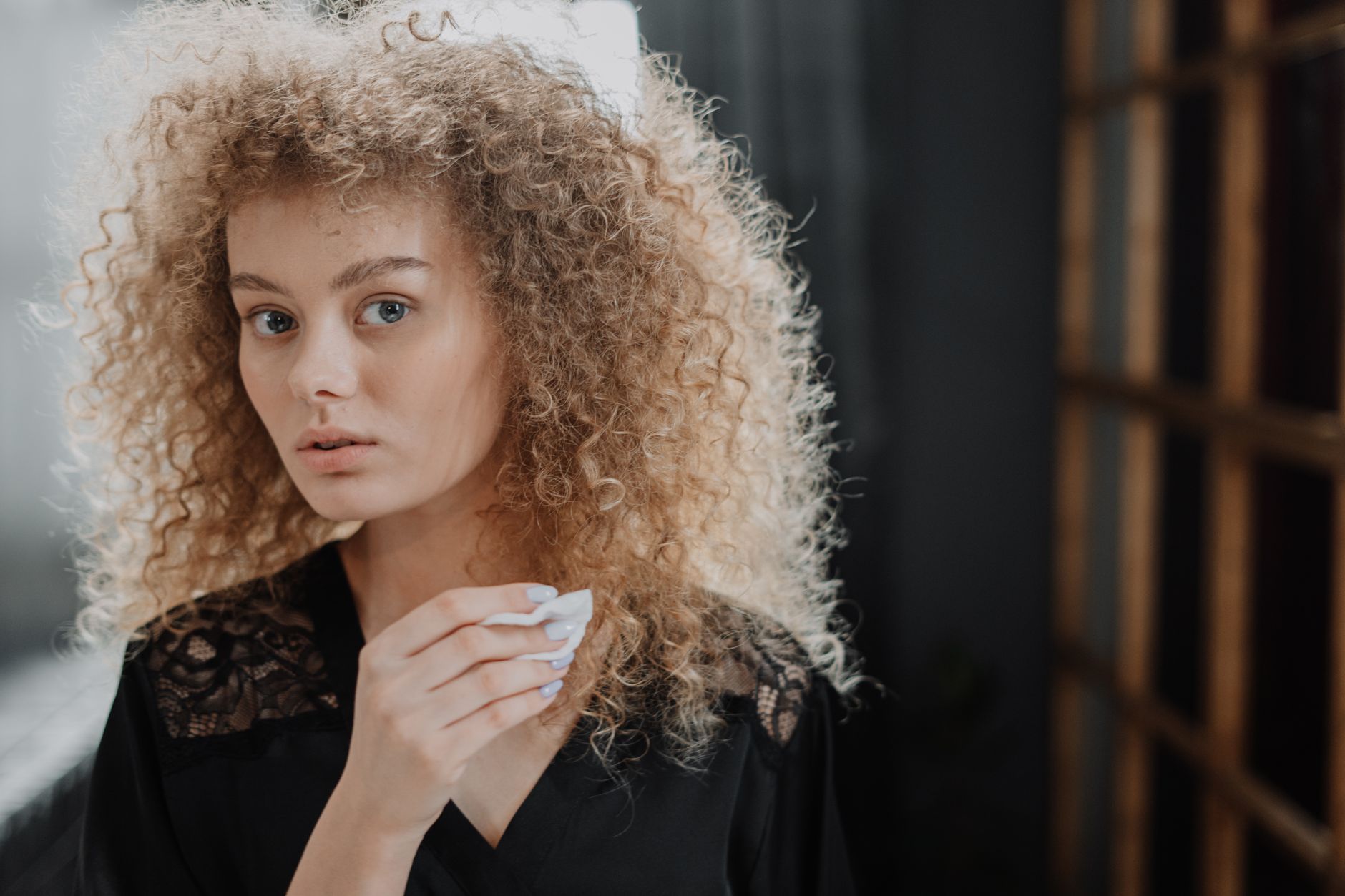 A young woman with curly hair using a cotton pad during her beauty routine indoors.