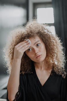 A young woman with curly hair cleanses her face with a cotton pad in a modern bathroom.