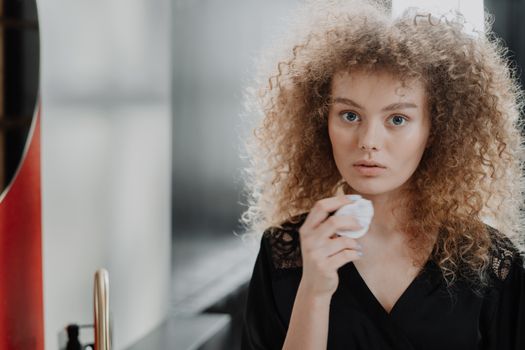 Young woman with curly hair applying beauty routine with cotton pad in a bathroom.