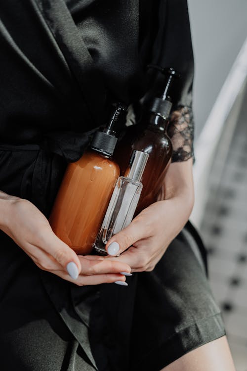 Person in Black Silk Robe Holding Glass Pump Bottles 