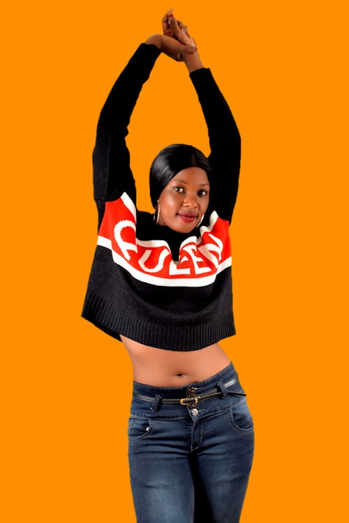 Positive young African American lady in trendy sweater with bare belly standing and raising hands in studio on orange background