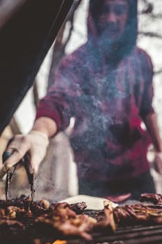 A man in a red sweater grills meat outside, surrounded by smoke, using tongs.