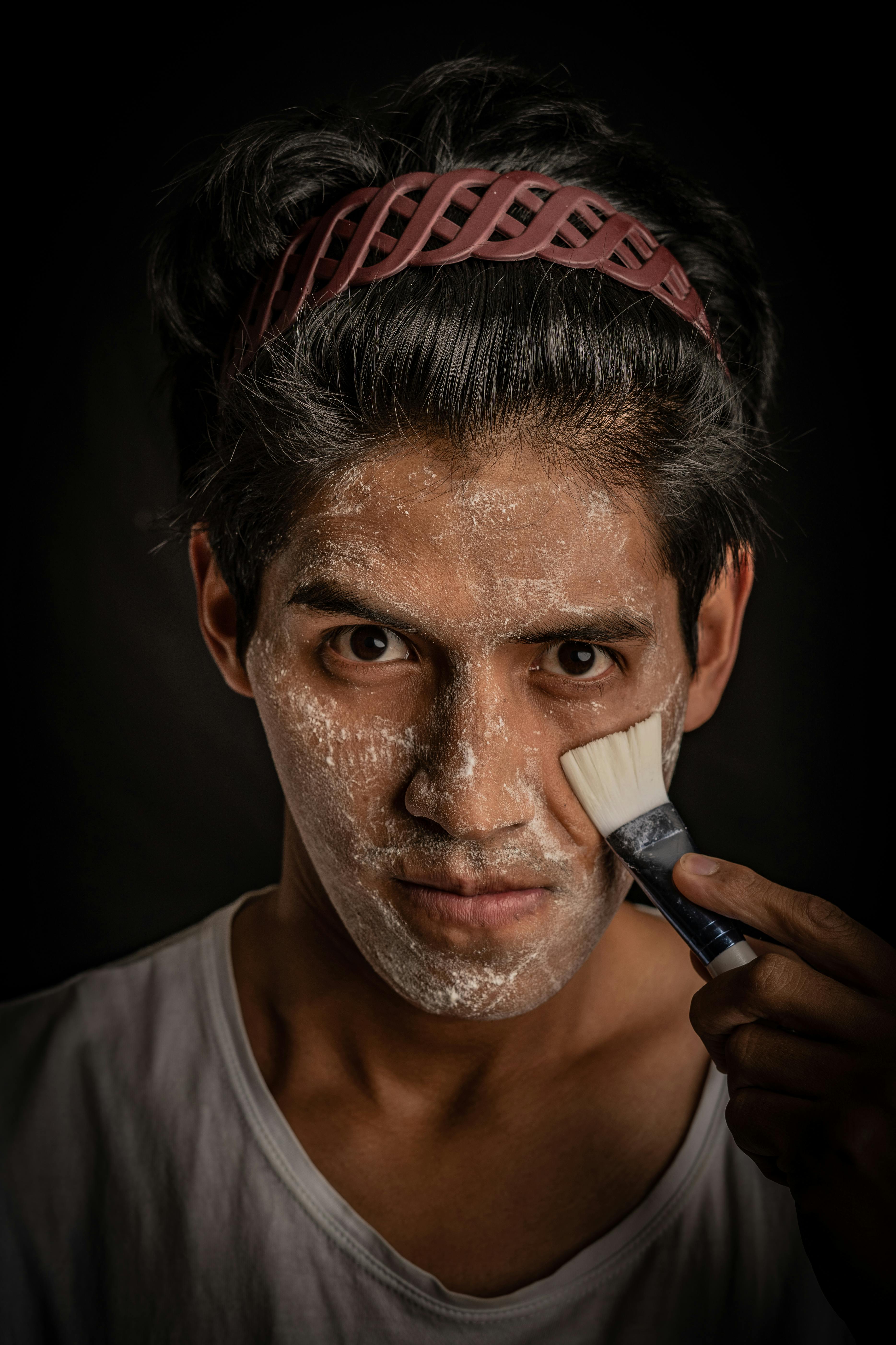 Free A dramatic studio portrait of a man applying white powder makeup with a brush against a black background. Stock Photo