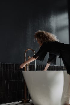 A young woman with curly hair preparing a bath in a dimly lit bathroom.