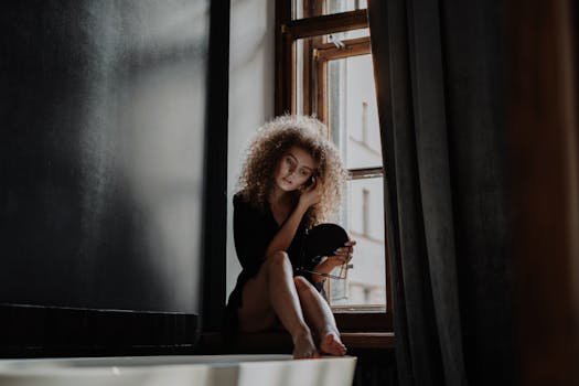 一位卷发的年轻女子沐浴在柔和的自然光线下，坐在窗前，手里拿着一面镜子