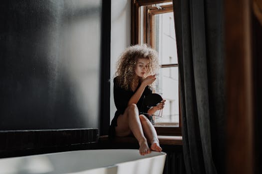 A young woman with curly hair sitting on a windowsill, holding a mirror.