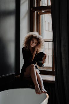 A young woman with curly hair sits by a window holding a mirror, capturing a moment of introspection.