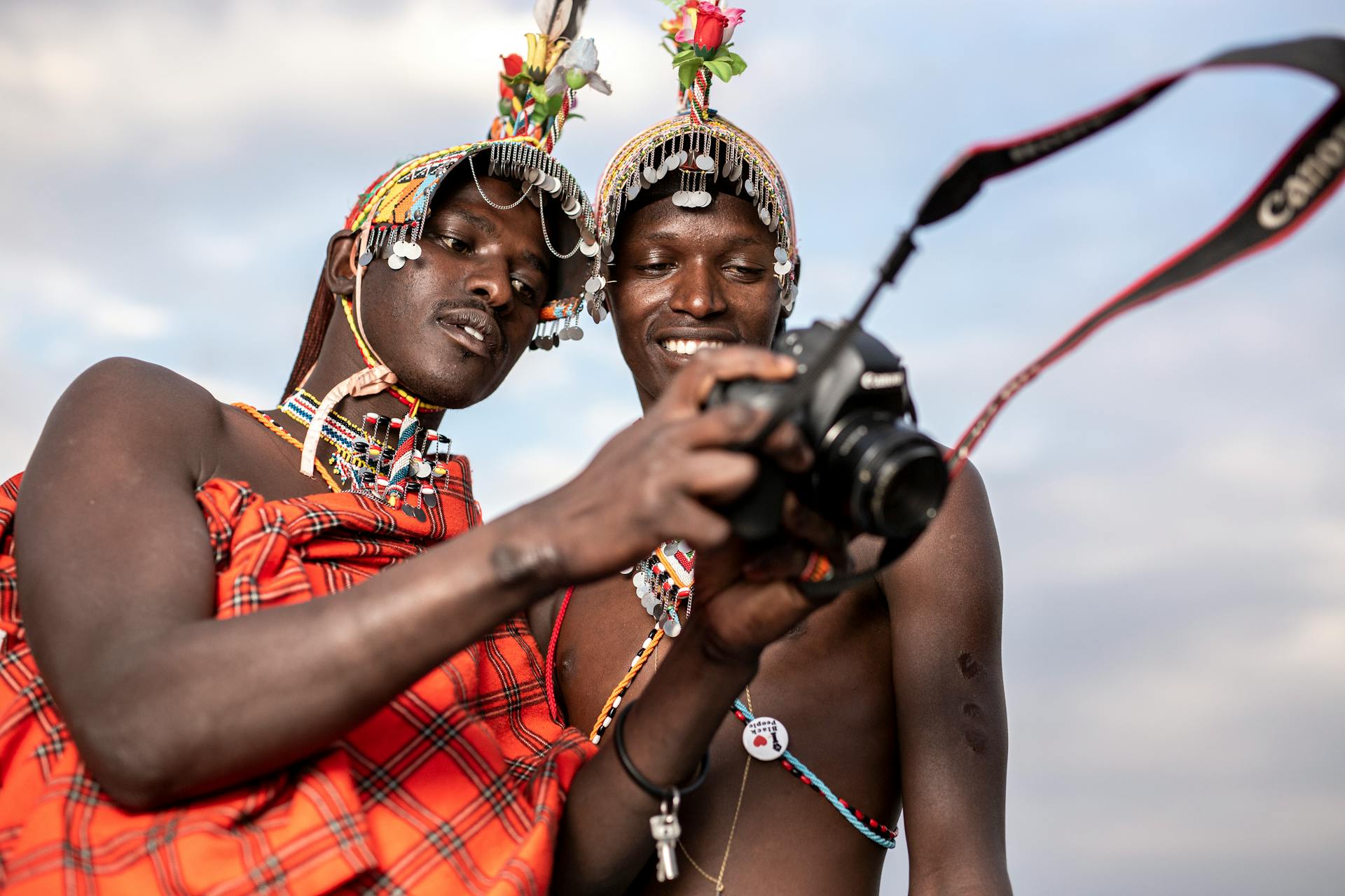 Two Kenyan men in traditional attire explore photography with a camera outdoors.