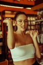 Provocative young female in trendy top playing with eyeglasses cord between teeth and looking at camera arrogantly while spending time in modern bookshop