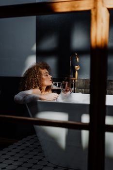 一位卷发女士正泡着泡泡浴，一边品着葡萄酒，一边放松身心