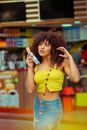Coquettish slim African American female in trendy wear standing with yummy pastry and gently touching curly hair against street food counter and looking away dreamily