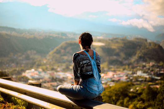 A woman sits on a guardrail enjoying a breathtaking mountain view.