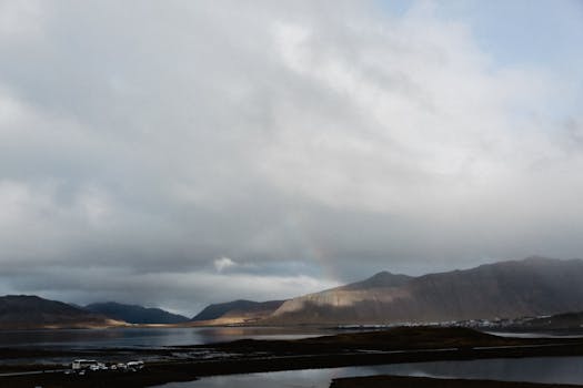 Scenic view of mountains and lake beneath a cloudy sky with a subtle rainbow.