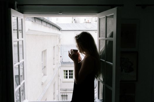 Silhouette of a woman sipping coffee by an open window with a city view.