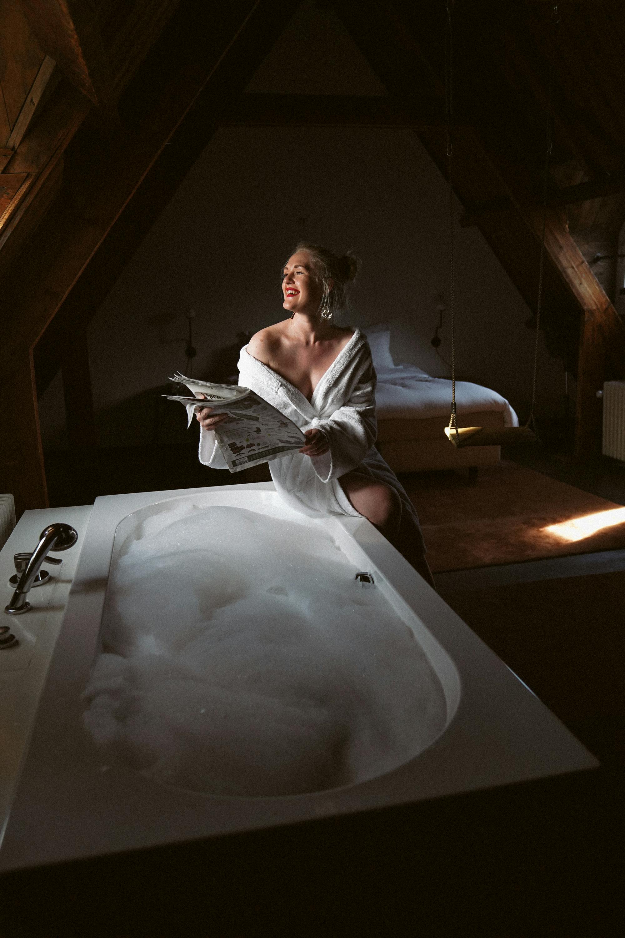 A woman enjoys a relaxing morning reading a newspaper next to a warm, bubbly bath in a rustic attic bedroom.