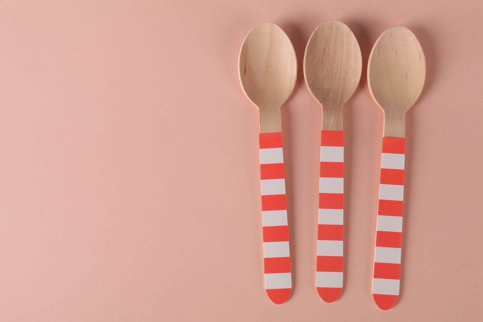 Three eco-friendly wooden spoons with striped handles on a minimalist pink background.