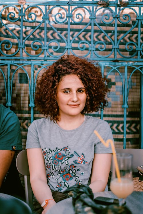 Free Positive young female with curly hair smiling and sitting at table with glass of cocktail Stock Photo