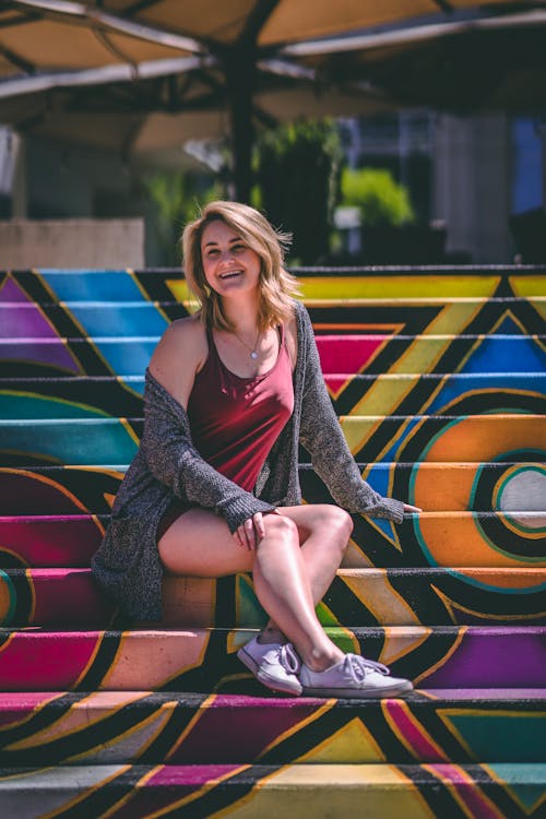 Full body of cheerful female traveler with blonde hair in comfortable wear looking away with bright smile while resting on vibrant stair painted with graffiti in sunlight