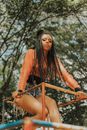 Low angle of serious young slim ethnic woman with bunch and braids looking at camera while resting on old shabby horizontal bar in park