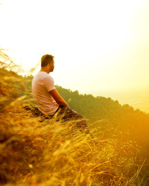 Free A solitary man sits on a grassy hill, watching the sunset during golden hour, creating a serene and introspective mood. Stock Photo