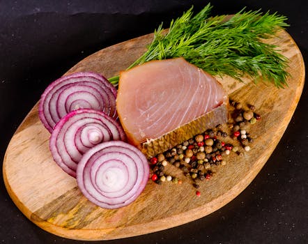 Close-up of sliced onion, dill, and fish fillet with mixed peppercorns on a wooden board.