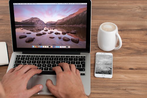 Person Using Macbook Pro on Brown Wooden Table Near Silver Iphone 6