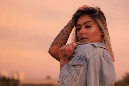 Free Serious young female with tattoos on arms in denim coat on shoulders looking at camera at sunset Stock Photo