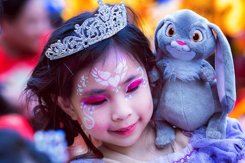Portrait of girl with crown on head and beautiful body art on face looking down with smile while holding toy on shoulder on blurred background