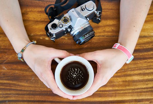Hands holding a cup of coffee beside a vintage camera on a wooden table.