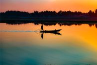 Back view of unrecognizable person fishing on traditional boat while floating on calm lake reflecting amazing colorful sunset sky