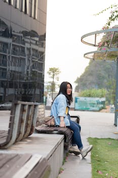 Young woman sitting casually on a bench outdoors with a modern cityscape background.