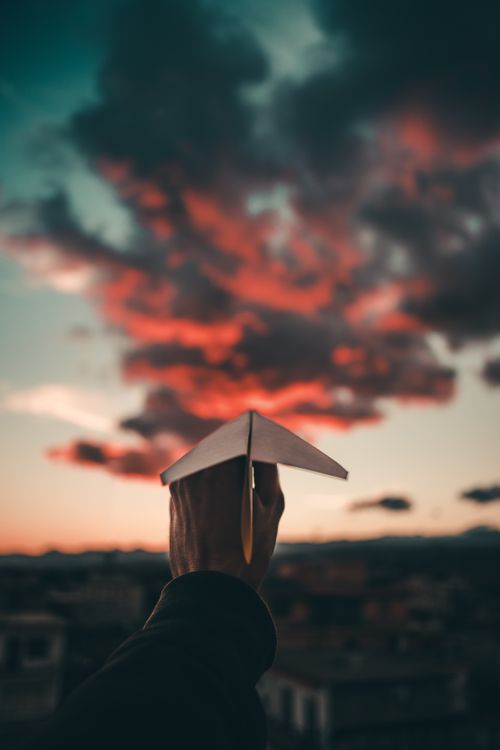 Free A person holds a paper plane against a striking sunset sky, capturing a whimsical moment. Stock Photo