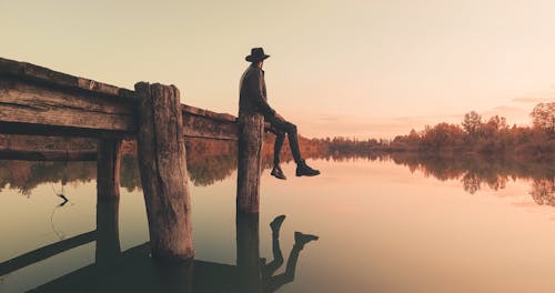 Schattenbild Des Mannes, Der Auf Holz Log In Wasser Während Des Sonnenuntergangs Sitzt