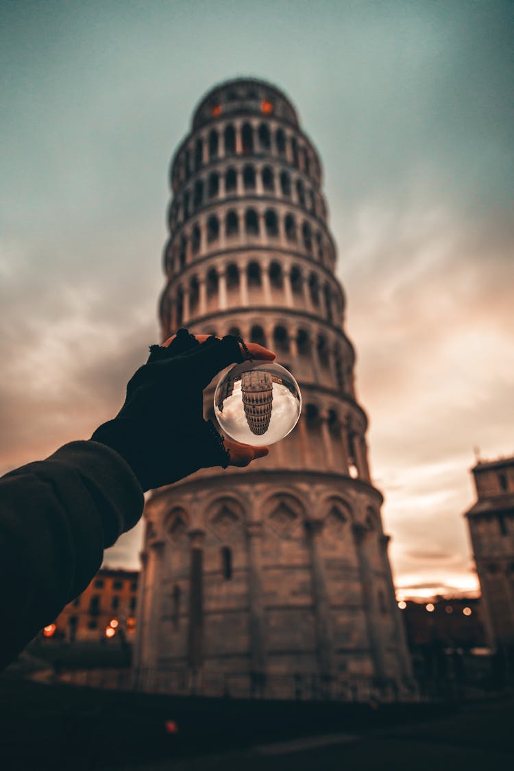 Hand Holding Glass Orb In Front Of Leaning Tower Of Pisa In Italy