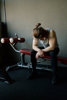 A male bodybuilder resting on a gym bench, looking exhausted after a workout session indoors.