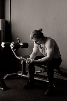 A man in a gym takes a pause during his workout, seated on a bench, showing muscular build.