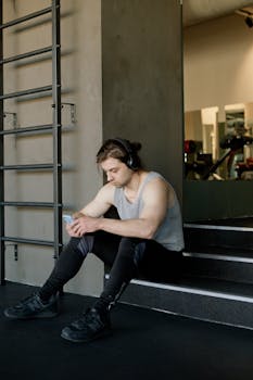 Young man in gym attire sitting on stairs with headphones and smartphone.