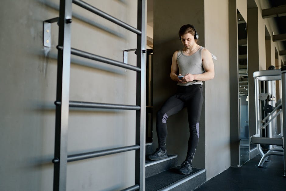 Man in gym standing on stairs, using smartphone while wearing headphones, dressed in sport wear.