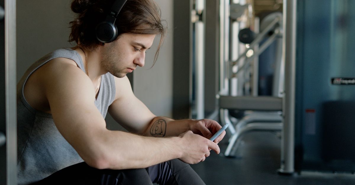 A young man wearing headphones sits in a gym, focused on texting with his smartphone.