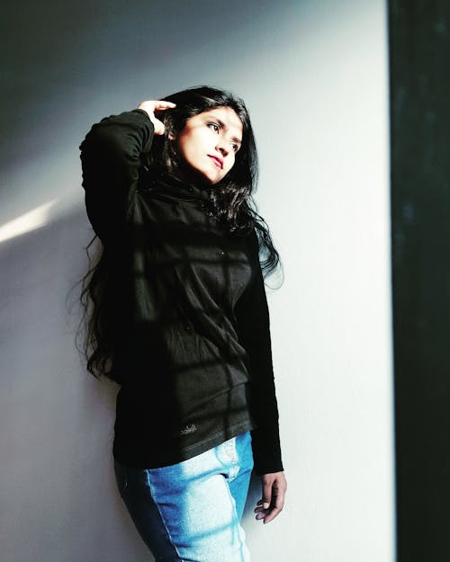 Pensive young ethnic female with long hair wearing denim with black hoodie looking away standing near white wall