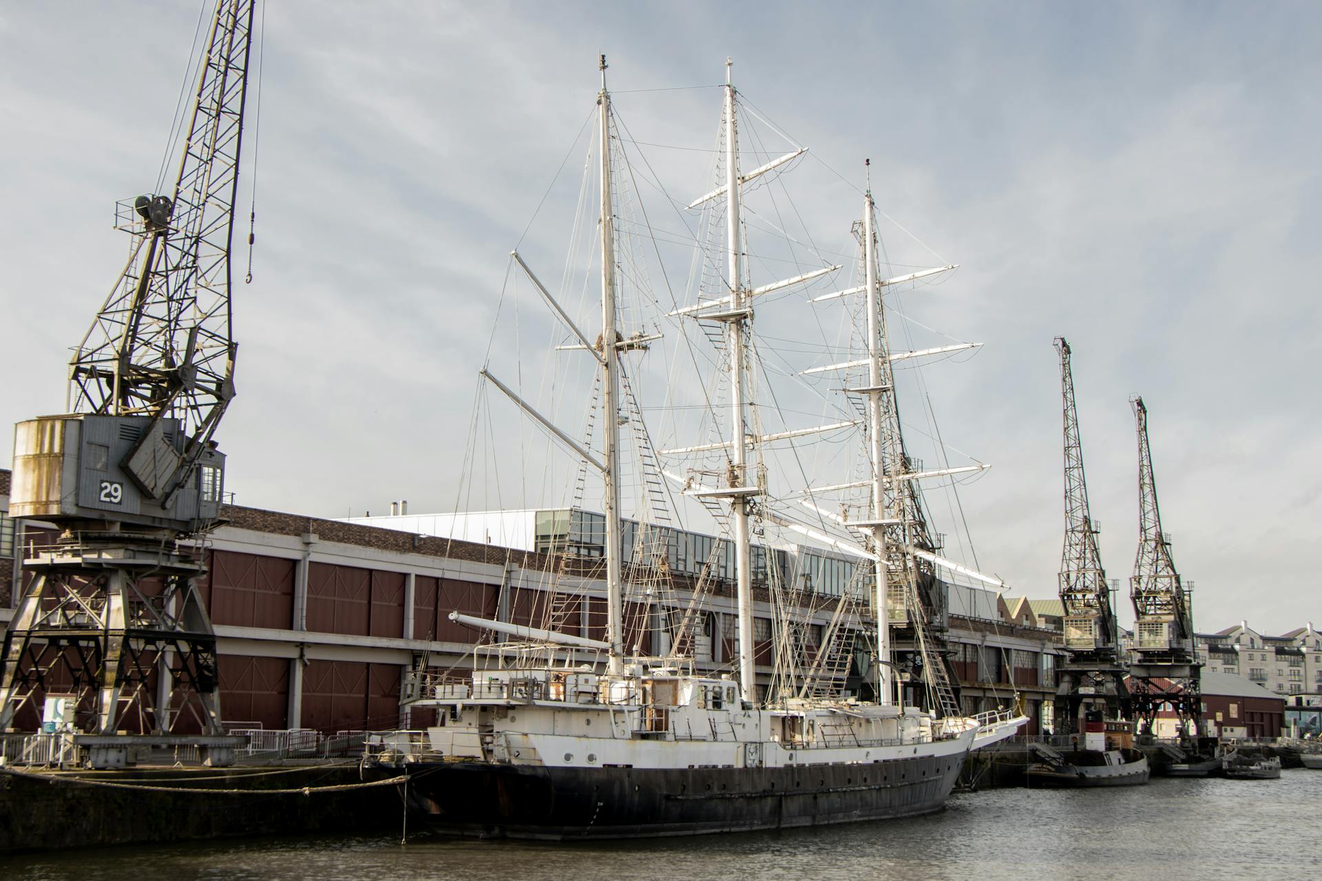 A vintage sailboat docked at an industrial harbor with surrounding cranes and buildings.