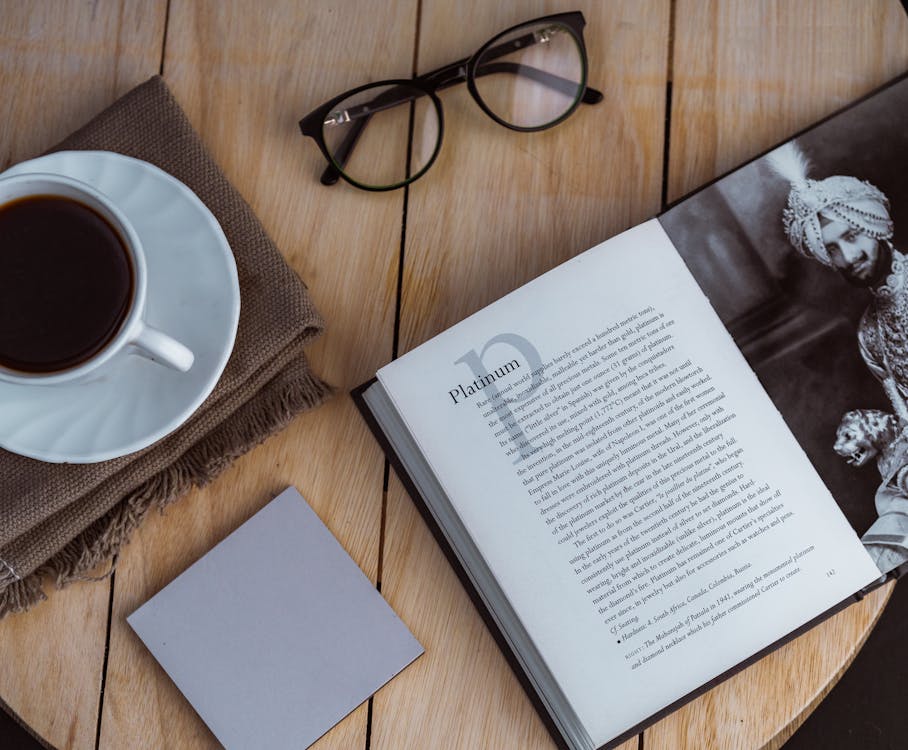 Opened book and sticky note placed on table with coffee cup and eyeglasses